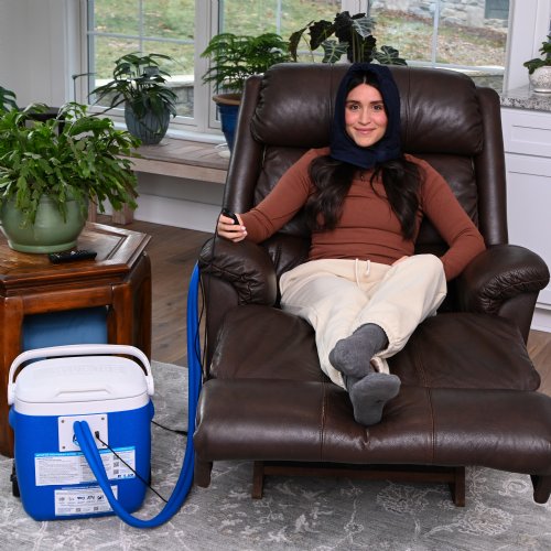 A young woman is standing upright wearing an Active Ice3.0 Cold Water Therapy Head Cap System the Head Cap is connected to the tubing of a 15 quart reservoir the reservoir's handle is fully extended 